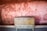 A vintage wooden cabinet with two doors is placed against a textured wall with a faded reddish hue. The wall, showing signs of wear, has a rough brick pattern and sporadic holes. The cabinet's surface is slightly worn, contributing to a rustic atmosphere.