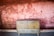 A vintage wooden cabinet with two doors is placed against a textured wall with a faded reddish hue. The wall, showing signs of wear, has a rough brick pattern and sporadic holes. The cabinet's surface is slightly worn, contributing to a rustic atmosphere.