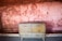 A vintage wooden cabinet with two doors is placed against a textured wall with a faded reddish hue. The wall, showing signs of wear, has a rough brick pattern and sporadic holes. The cabinet's surface is slightly worn, contributing to a rustic atmosphere.