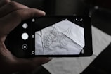 A person is holding a smartphone over a crumpled sheet of paper with a hand-drawn scorpion sketch on it. The phone's camera interface is visible on the screen, capturing the drawing.