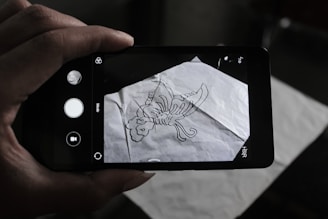 A person is holding a smartphone over a crumpled sheet of paper with a hand-drawn scorpion sketch on it. The phone's camera interface is visible on the screen, capturing the drawing.