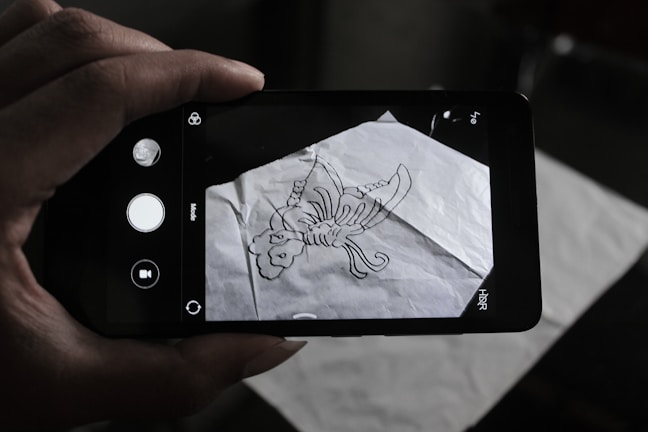 A person is holding a smartphone over a crumpled sheet of paper with a hand-drawn scorpion sketch on it. The phone's camera interface is visible on the screen, capturing the drawing.