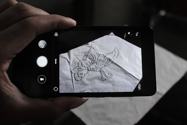 A person is holding a smartphone over a crumpled sheet of paper with a hand-drawn scorpion sketch on it. The phone's camera interface is visible on the screen, capturing the drawing.