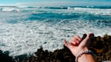 A scenic view of a vast ocean with waves crashing against dark rocks in the foreground. A hand, adorned with a beaded bracelet, is extended in the foreground, gesturing a shaka sign. The sky is mostly clear with a few clouds.