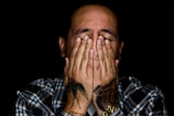 A person with tattoos on their hands covering their face, wearing a plaid shirt. The background is dark, highlighting the subject's distressed or contemplative expression.