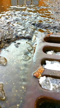 A close-up view of a street drain partially covered with melting ice and water. The surface around the drain is wet and slightly muddy, with small stones and debris scattered about. The lighting creates reflections and highlights on the water's surface.