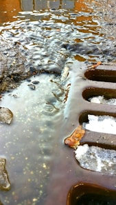 A close-up view of a street drain partially covered with melting ice and water. The surface around the drain is wet and slightly muddy, with small stones and debris scattered about. The lighting creates reflections and highlights on the water's surface.