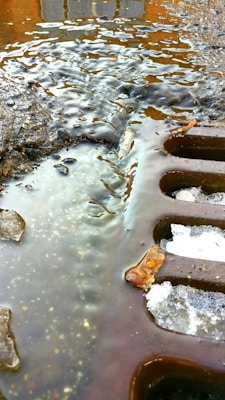 A close-up view of a street drain partially covered with melting ice and water. The surface around the drain is wet and slightly muddy, with small stones and debris scattered about. The lighting creates reflections and highlights on the water's surface.
