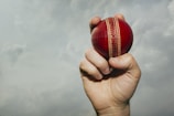 A hand is gripping a red cricket ball against a backdrop of a cloudy sky. The ball is prominently displayed, revealing its stitched seam.