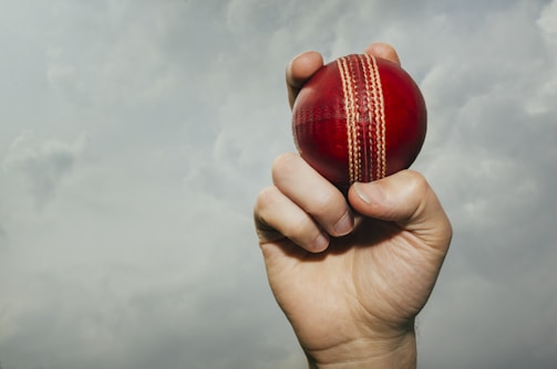 A hand is gripping a red cricket ball against a backdrop of a cloudy sky. The ball is prominently displayed, revealing its stitched seam.