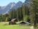 A serene landscape featuring a rustic wooden cabin and a barn nestled in a lush green meadow. Towering pine trees surround the area, and majestic mountains rise in the background under a clear blue sky.