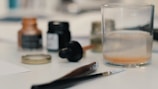 A close-up view of a workspace featuring art supplies. There is a glass filled with water, an open bottle of ink, and several paintbrushes laying on the table. The scene includes a mix of blurred objects in the background, giving a sense of creative activity.