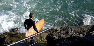A person in a wetsuit stands at the edge of rocky stairs holding an orange surfboard, looking at the choppy sea. The ocean waves crash against the rocks, creating a dynamic and adventurous atmosphere.