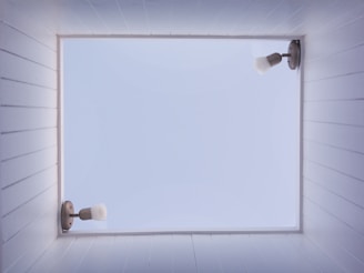 A square skylight framed by white wooden planks on all sides reveals a clear blue sky. Two wall-mounted light fixtures are on opposite sides of the skylight frame, featuring metal mounts and white bulb covers pointing away from the center.