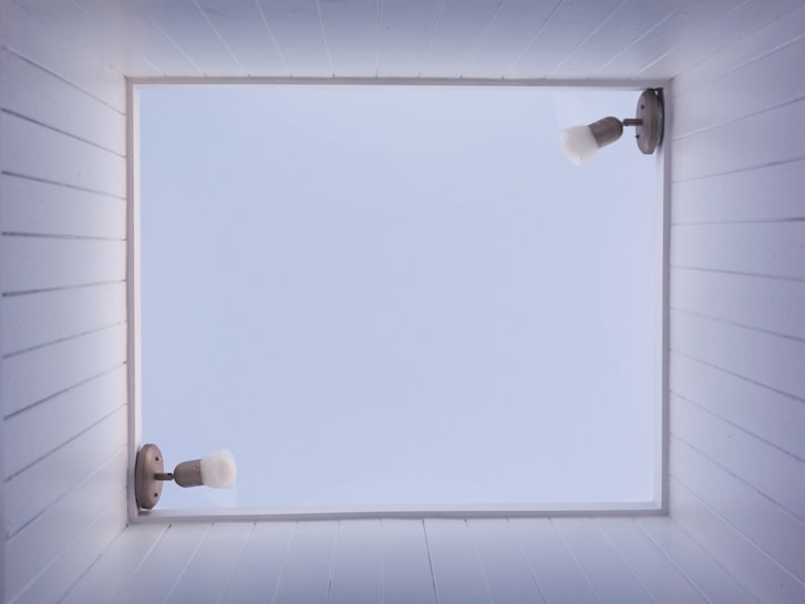 A square skylight framed by white wooden planks on all sides reveals a clear blue sky. Two wall-mounted light fixtures are on opposite sides of the skylight frame, featuring metal mounts and white bulb covers pointing away from the center.