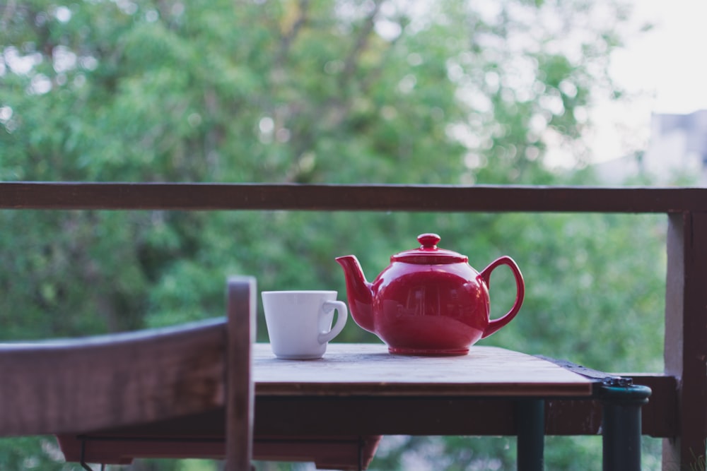 Foto de foco seletivo de bule de cerâmica vermelha e xícara de chá de cerâmica branca na mesa de madeira marrom perto da árvore durante o dia