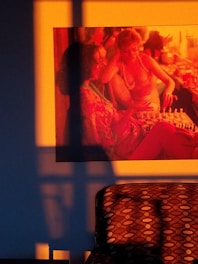 A warm-toned image features a painting or print of two women playing chess, surrounded by vibrant orange and red hues. The artwork is mounted on a wall with an intricate pattern or texture that reflects light and shadow. Below, a patterned chair is partially visible, with a polka dot design, adding to the artistic ambiance.