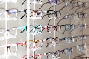 Multiple pairs of eyeglasses arranged neatly on a display rack. The eyeglasses come in various colors and frame styles, and they are evenly spaced and organized in rows.
