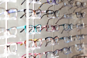 Multiple pairs of eyeglasses arranged neatly on a display rack. The eyeglasses come in various colors and frame styles, and they are evenly spaced and organized in rows.