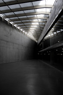 A modern architectural space featuring tall, gray concrete walls and a ceiling with glass panels allowing natural light to filter in. The perspective leads towards a vanishing point, creating a sense of depth. The space is empty and minimalistic, with a smooth, reflective floor.