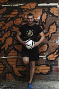 A man is leaning against a graffiti-covered brick wall while holding a soccer ball. He is dressed in a black t-shirt and shorts, and wearing blue athletic shoes. The background features bold, abstract black and orange graffiti art on the wall.