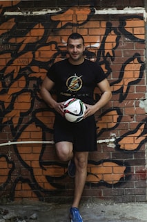 A man is leaning against a graffiti-covered brick wall while holding a soccer ball. He is dressed in a black t-shirt and shorts, and wearing blue athletic shoes. The background features bold, abstract black and orange graffiti art on the wall.