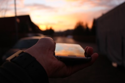 A hand holding a smartphone outdoors with a scenic sunset reflected on the screen.