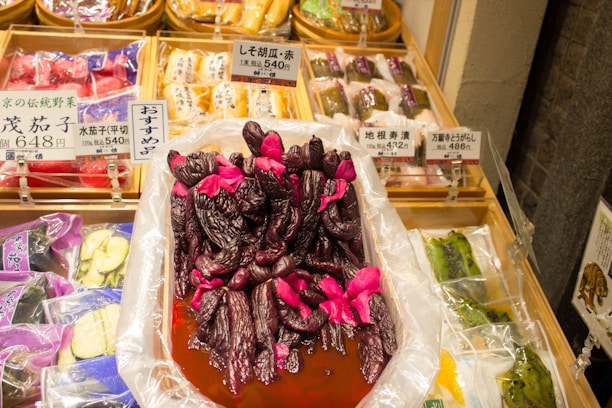 A variety of pickled vegetables are displayed in a market setting, featuring a prominent tray of deep purple pickled eggplants with bright pink accents being the focal point. Surrounding this tray, there are neatly arranged packages of other pickled vegetables, each with a distinct label displaying prices and descriptions in Japanese.