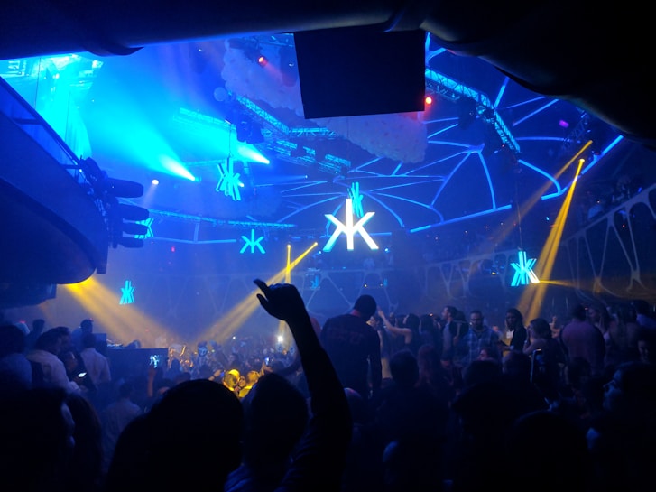 A crowded nightclub scene with a large number of people dancing and socializing under vibrant blue and yellow lights. The club features a modern design with geometric patterns on the ceiling and an illuminated logo at the center. The atmosphere is lively and energetic.