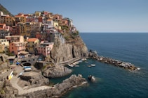 A picturesque coastal village with colorful buildings perched on a rocky cliff overlooking the sea. Several small boats are moored in the turquoise harbor below. The area is bustling with people and activity, with pathways winding through the charming townscape.
