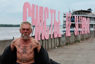 An older man with tattoos is standing by a waterfront smoking, with large pink letters forming a sign in the background, next to a docked ship.