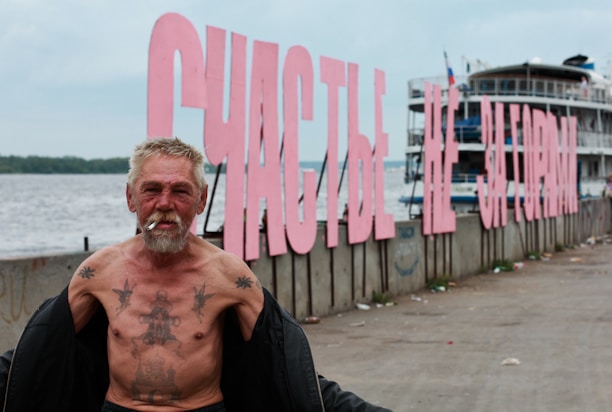 An older man with tattoos is standing by a waterfront smoking, with large pink letters forming a sign in the background, next to a docked ship.
