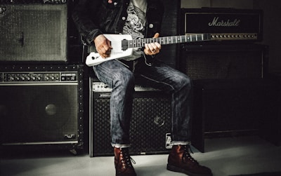 A person in casual attire is holding and playing an electric guitar. They are seated on a large speaker, surrounded by several amplifiers, including a prominent Marshall amp. The background is dimly lit, highlighting the vintage musical equipment.