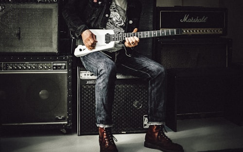 man wearing a black jacket, white shirt, blue jeans, and brown boots, playing a white electric guitar while sitting on an amp