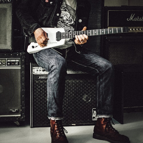 man wearing a black jacket, white shirt, blue jeans, and brown boots, playing a white electric guitar while sitting on an amp