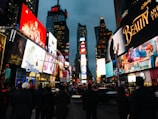 An urban scene filled with bright, colorful billboards and advertisements, likely in a busy city area. Crowds of people gather on the street near lit-up signage. There's a prominent display of movie advertisements and corporate logos on the buildings.