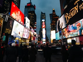 An urban scene filled with bright, colorful billboards and advertisements, likely in a busy city area. Crowds of people gather on the street near lit-up signage. There's a prominent display of movie advertisements and corporate logos on the buildings.