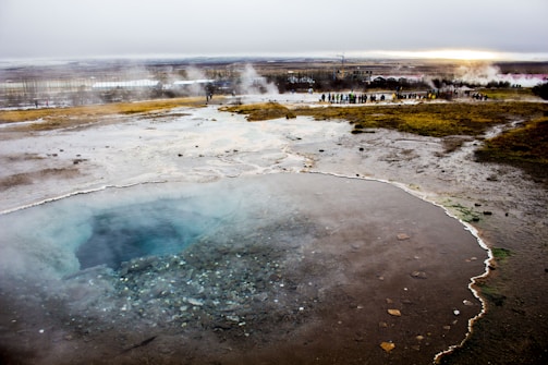 A large geothermal pool is steaming in the foreground, with clear, blue water surrounded by rocky terrain. A line of people is visible in the background, walking along a path in a wide, open landscape with patches of vegetation. The sky is overcast, and steam is rising from several spots across the area.