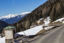 A picturesque mountainous landscape with snow-covered peaks in the background and a winding road in the foreground. A small building with a green roof sits on the left side near a snow-covered area, surrounded by a mix of evergreen and deciduous trees.