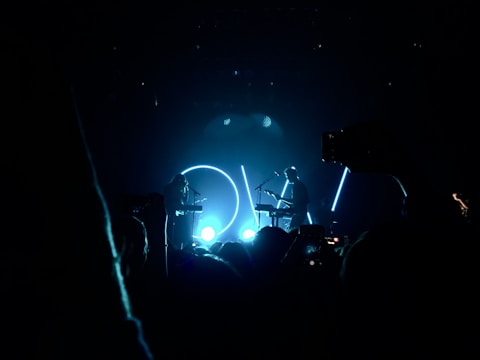 Silhouetted musicians perform on stage, illuminated by vibrant blue lights and circular backlighting. The crowd captures the moment with their smartphones, highlighting the concert atmosphere.