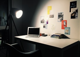 A dimly lit workspace features a modern desk lamp casting light on a minimalist white desk. A laptop sits open alongside a stack of books. Various art prints and motivational posters are arranged on the wall, adding color and personality to the space.
