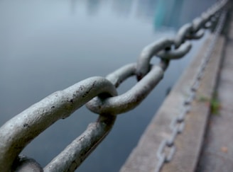 A metal chain stretches along the edge of a waterfront, with individual links of the chain in focus and the water out of focus in the background. The chain is attached to concrete surfaces, creating a sense of boundary or barrier.