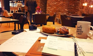 A cozy cafe setting with wooden tables and chairs, featuring a notebook and papers on a table with a pen, a large white mug with the phrase 'Freshly Baked, Every day', and a plate of pastries. In the background, people are seated and walking around, with warm lighting and a brick wall.