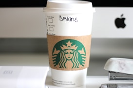 A white takeaway coffee cup with a brown cardboard sleeve featuring the green siren logo is placed on a desk. The cup has a handwritten name, possibly indicating it is from a popular coffee chain. In the background is the edge of a computer screen with a visible logo, along with an out-of-focus stapler and other desk items.