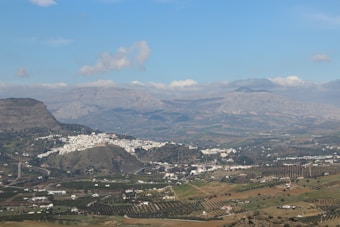 A picturesque landscape featuring a sprawling valley with a whitewashed village nestled on a hillside. The scene is framed by distant mountain ranges under a clear blue sky with scattered clouds. Agricultural fields with rows of trees, likely olive groves, are visible in the foreground, interspersed with roads and smaller clusters of buildings.
