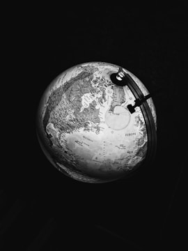 A monochrome image of a lit globe displaying various geographical locations and labels. The focus is primarily on the Northern Hemisphere, showing parts of Europe and the North Atlantic Ocean. The lighting creates strong contrasts between the illuminated map and the surrounding darkness.