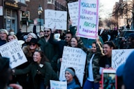 A diverse group of people are gathered on a street holding various protest signs. The signs convey messages about feminism, anti-racism, and unity. The participants are smiling and appear engaged with the event, suggesting a peaceful demonstration. The background includes urban elements like buildings and street lamps.