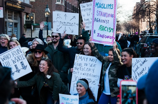 A diverse group of people are gathered on a street holding various protest signs. The signs convey messages about feminism, anti-racism, and unity. The participants are smiling and appear engaged with the event, suggesting a peaceful demonstration. The background includes urban elements like buildings and street lamps.