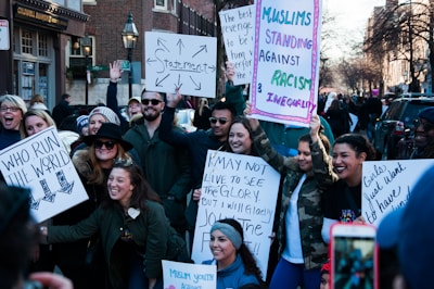 A diverse group of people are gathered on a street holding various protest signs. The signs convey messages about feminism, anti-racism, and unity. The participants are smiling and appear engaged with the event, suggesting a peaceful demonstration. The background includes urban elements like buildings and street lamps.