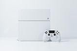 A white gaming console standing upright with a matching white controller placed in front of it. The background is a light, neutral color, accentuating the sleek and modern design of the console.